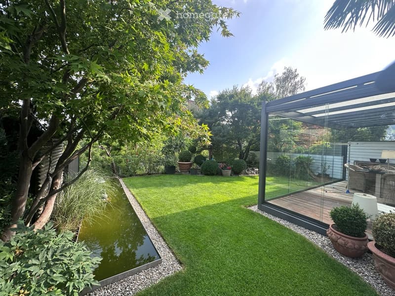 Well-maintained garden with a lawn, a small water feature, potted plants, and a modern glass-walled extension.