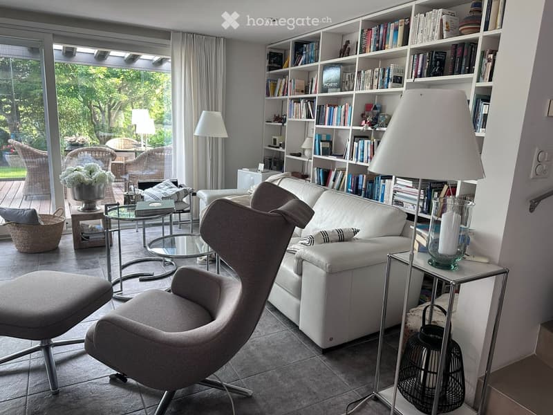 Modern living room with a white leather sofa, armchair, glass coffee table, bookshelf filled with books, and a large window with a view of the garden.