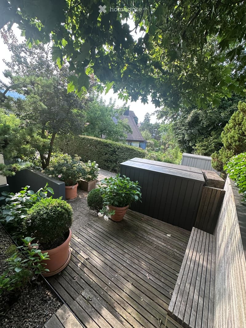 Wooden deck with potted plants, surrounded by trees and greenery, with a view of neighboring houses.