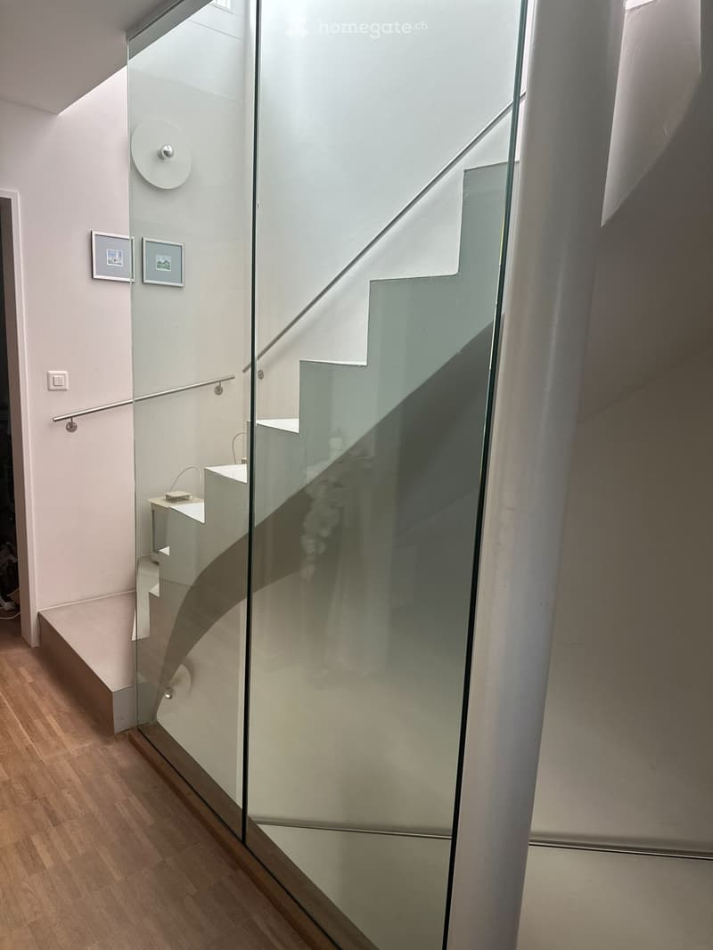 Modern hallway with wooden flooring, glass staircase, and white walls. Features a small table with a lamp and framed pictures on the wall.