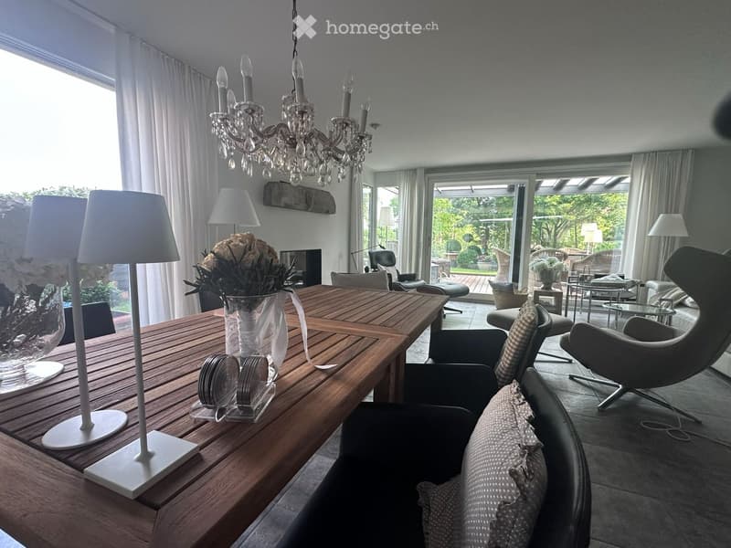 Modern living room with a wooden dining table, sofa, chairs, chandelier, lamps, and a view of the garden through large windows.