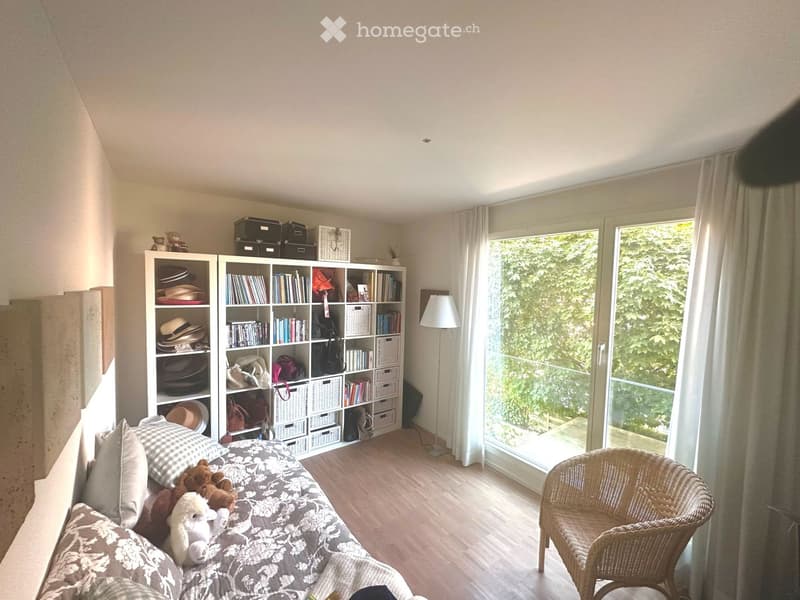 Bright bedroom with a daybed, bookshelf, and large window with a view of greenery. The room features a wicker chair and decorative stuffed animals on the bed.