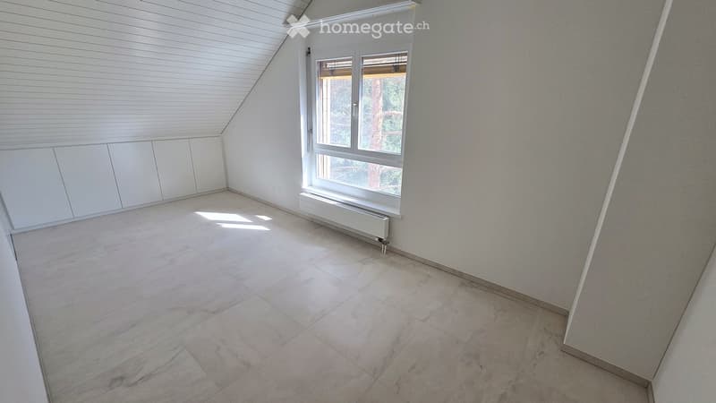 Empty attic room with white walls, wooden slatted ceiling, large window, and tiled floor.