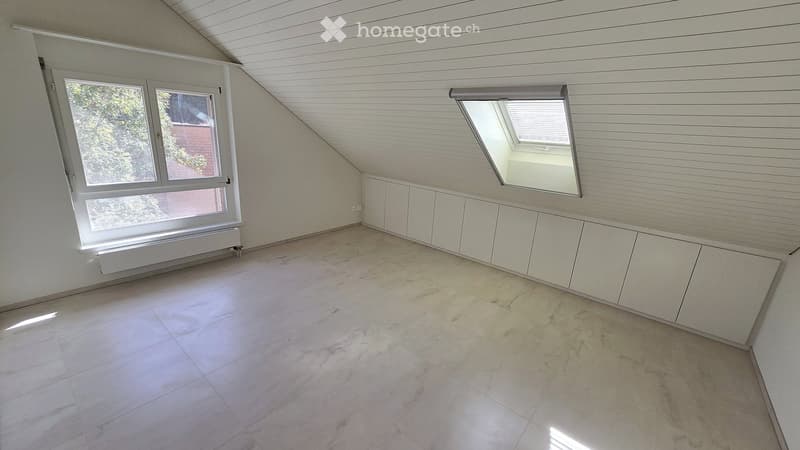 Empty attic room with white walls, tiled floor, large windows, and built-in white cabinets.