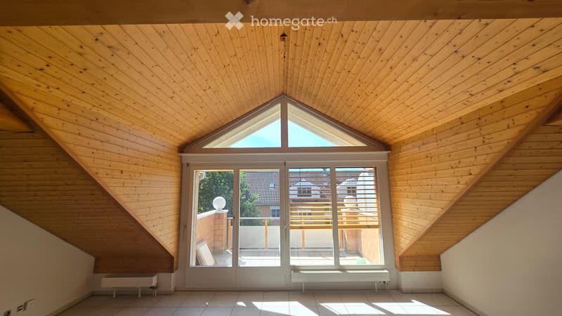 Bright, wooden-paneled attic room with skylight and large glass doors leading to a balcony. The space is empty, with only built-in features like the wooden ceiling and tiled floor visible.