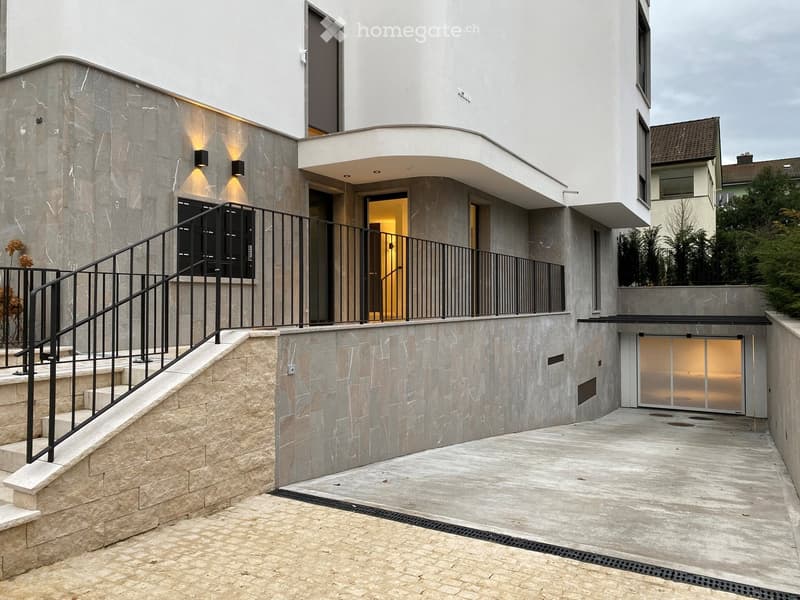 Modern two-story building with a raised ground floor, featuring a curved balcony, stone staircase, and illuminated entrance.