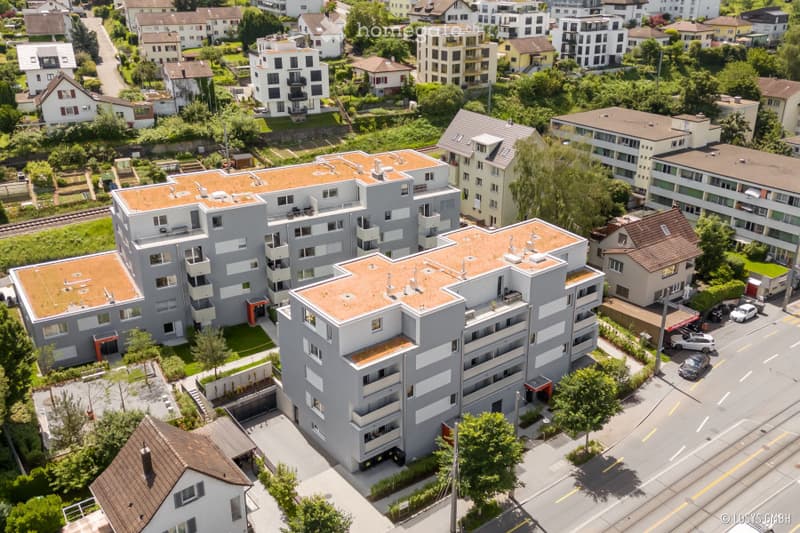 Aerial view of a modern multi-story residential building complex with grey facades, orange-tiled roofs, and surrounding greenery.