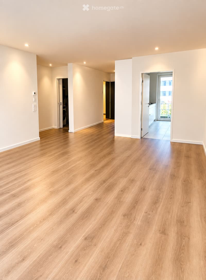 Empty room with light wooden flooring and white walls, featuring recessed lighting and open doorways leading to other rooms.