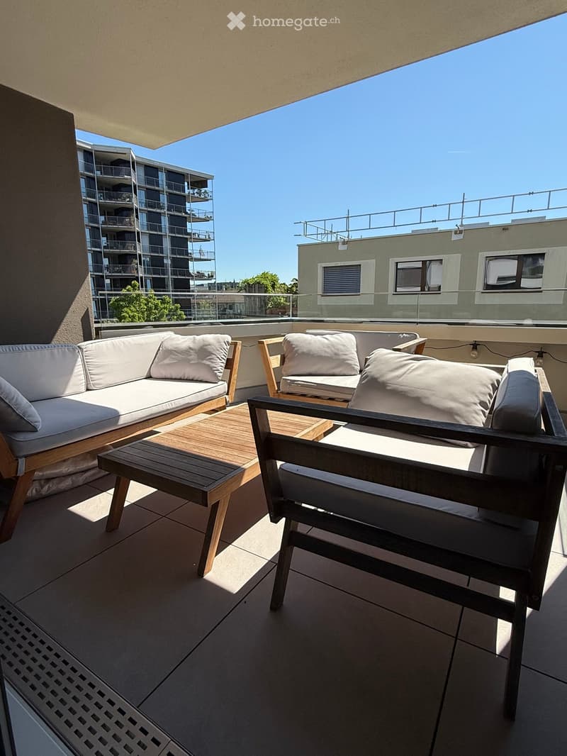 Modern rooftop terrace with a white sofa, wooden chairs, and a small wooden table, overlooking a cityscape with modern buildings.
