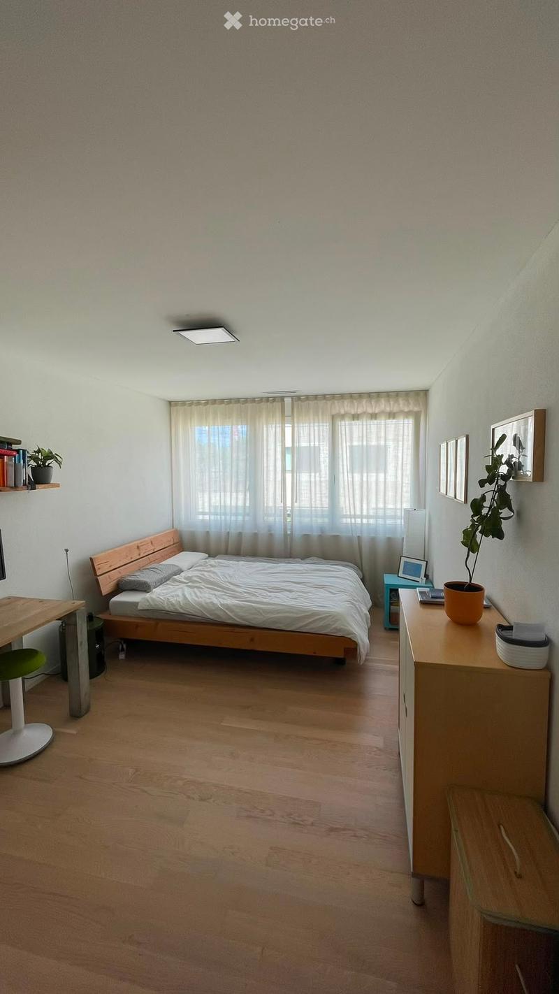 Cozy bedroom with wooden flooring, a bed with white bedding, a nightstand with a lamp, a desk with a chair, and a shelf with books and a plant.