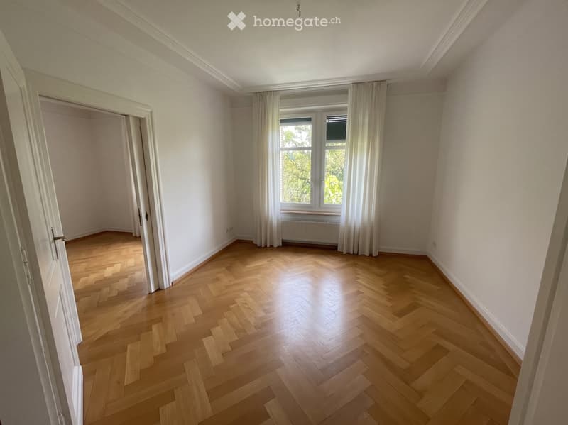Empty room with light wooden flooring, white walls, large window with white curtains, and a visible doorway leading to another room.