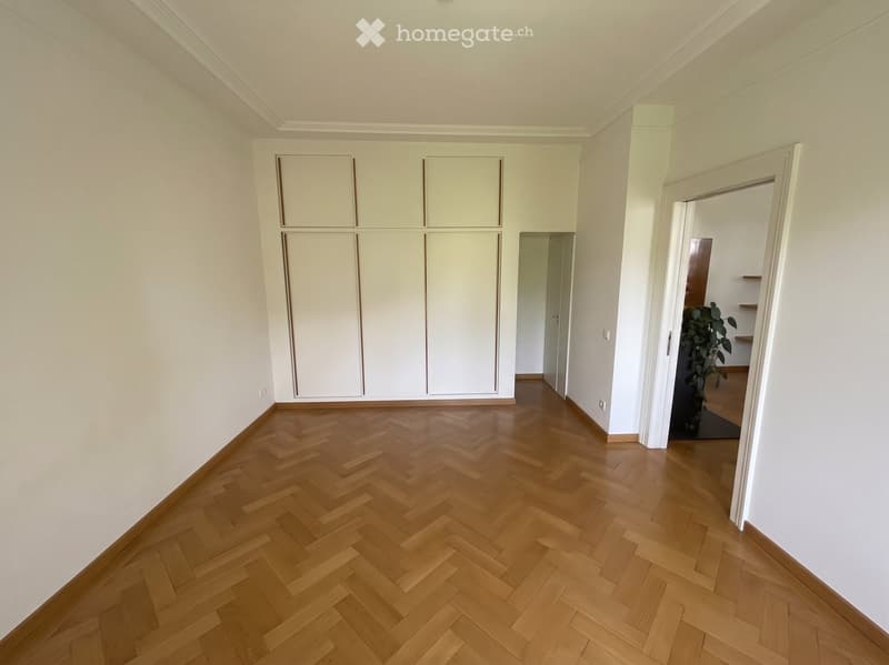 Empty room with wooden herringbone flooring, white walls, and built-in white closet units. A doorway leads to another room with a visible plant and shelves.
