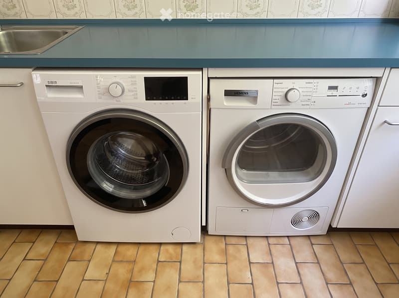 A bathroom with a washing machine and dryer installed side by side, featuring tiled flooring and light blue cabinets.
