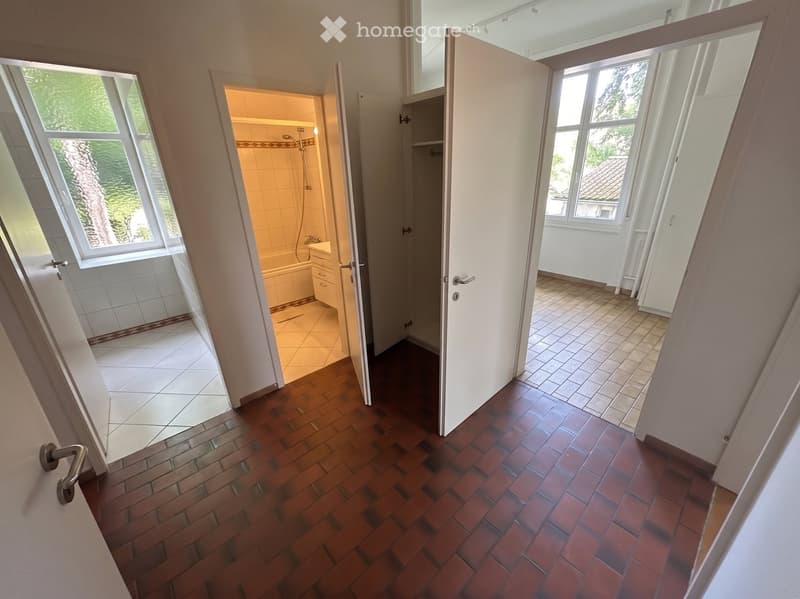 Hallway with wooden and tiled flooring, leading to a bathroom and another room with large windows and natural light.