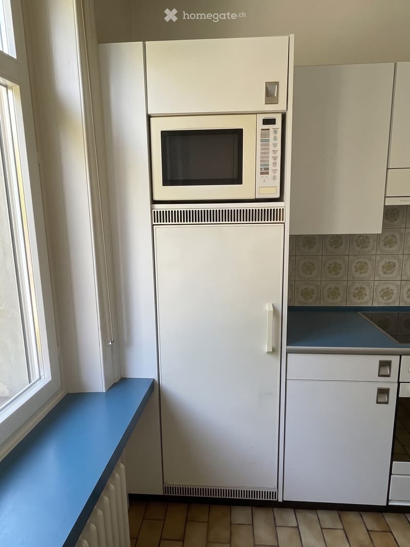 A compact kitchen with a white refrigerator, microwave, and cabinets. The floor is tiled, and there is a blue countertop near a window.