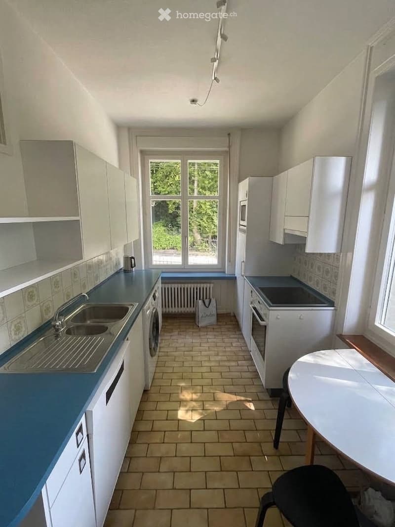 A modern kitchen with white cabinets, blue countertops, a sink, an oven, a refrigerator, a microwave, a small table, and a chair. The room has tiled flooring and large windows letting in natural light.