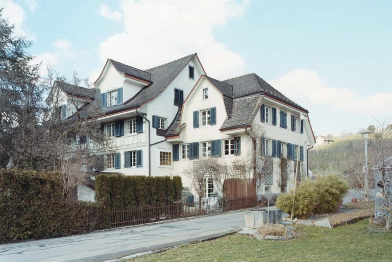 A two-story white house with blue shutters, multiple windows, and a sloped roof. The house is surrounded by a low hedge, a small garden area, and a paved driveway.