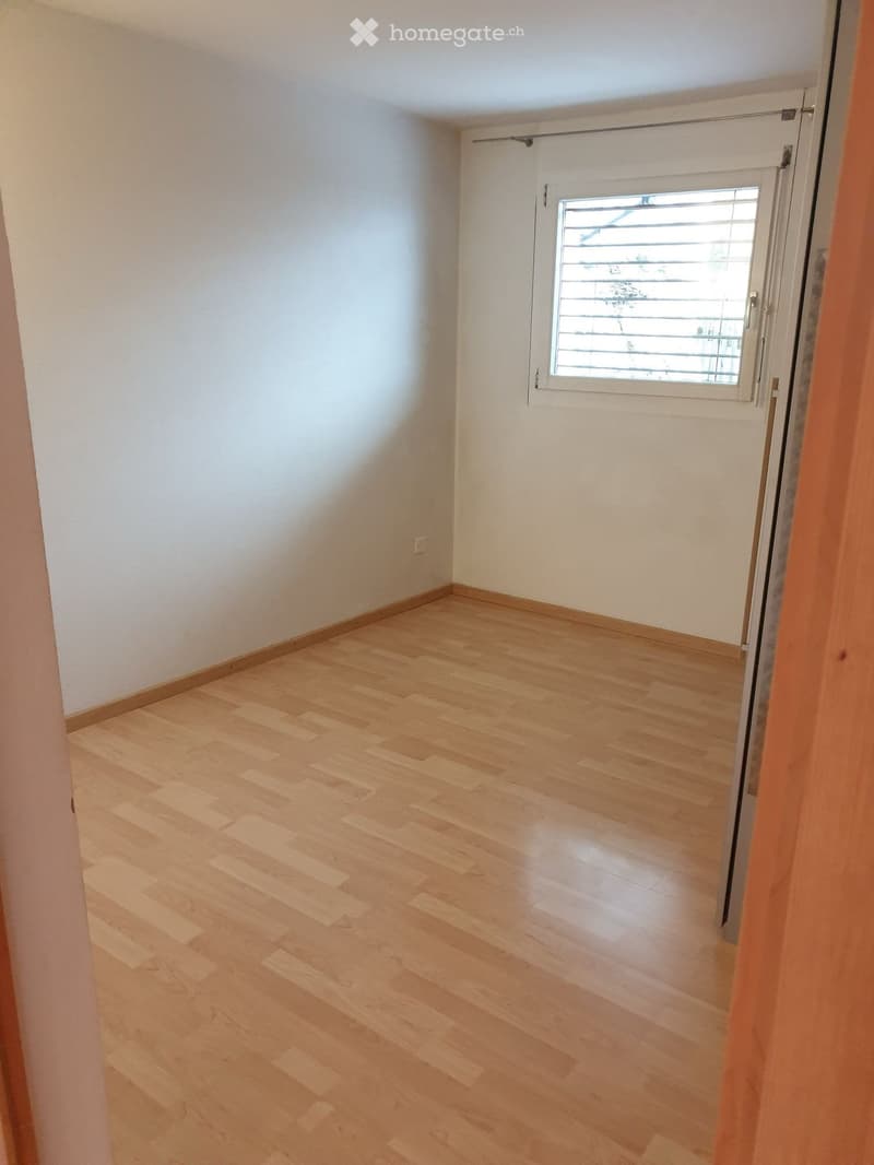 Empty room with light wooden flooring and white walls, featuring a window with blinds and a door frame visible.