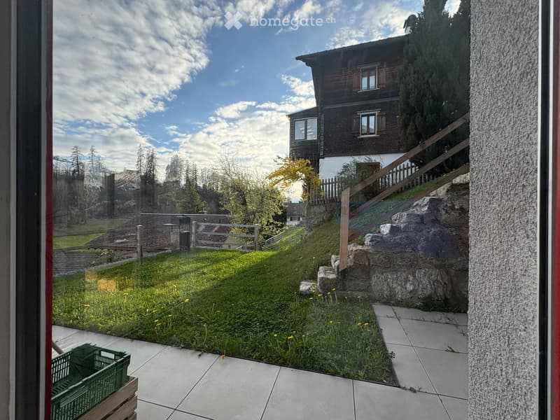 View from a balcony or terrace showing a grassy garden area with small yellow flowers, a wooden staircase leading up to a house, and a wooden fence. The house has a dark wooden facade with multiple windows and is surrounded by trees and a clear blue sky.