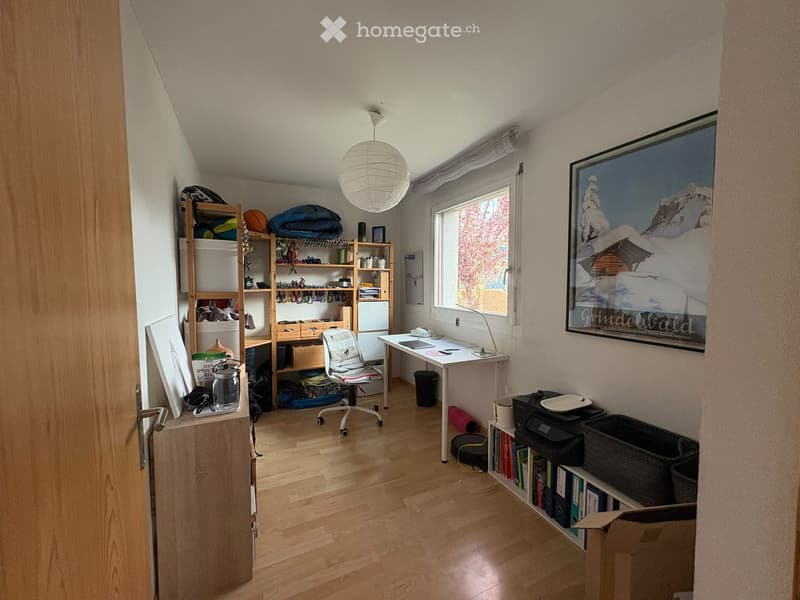 A well-lit room with wooden flooring, a large window, a desk with a chair, shelves filled with books and items, and a picture frame on the wall.
