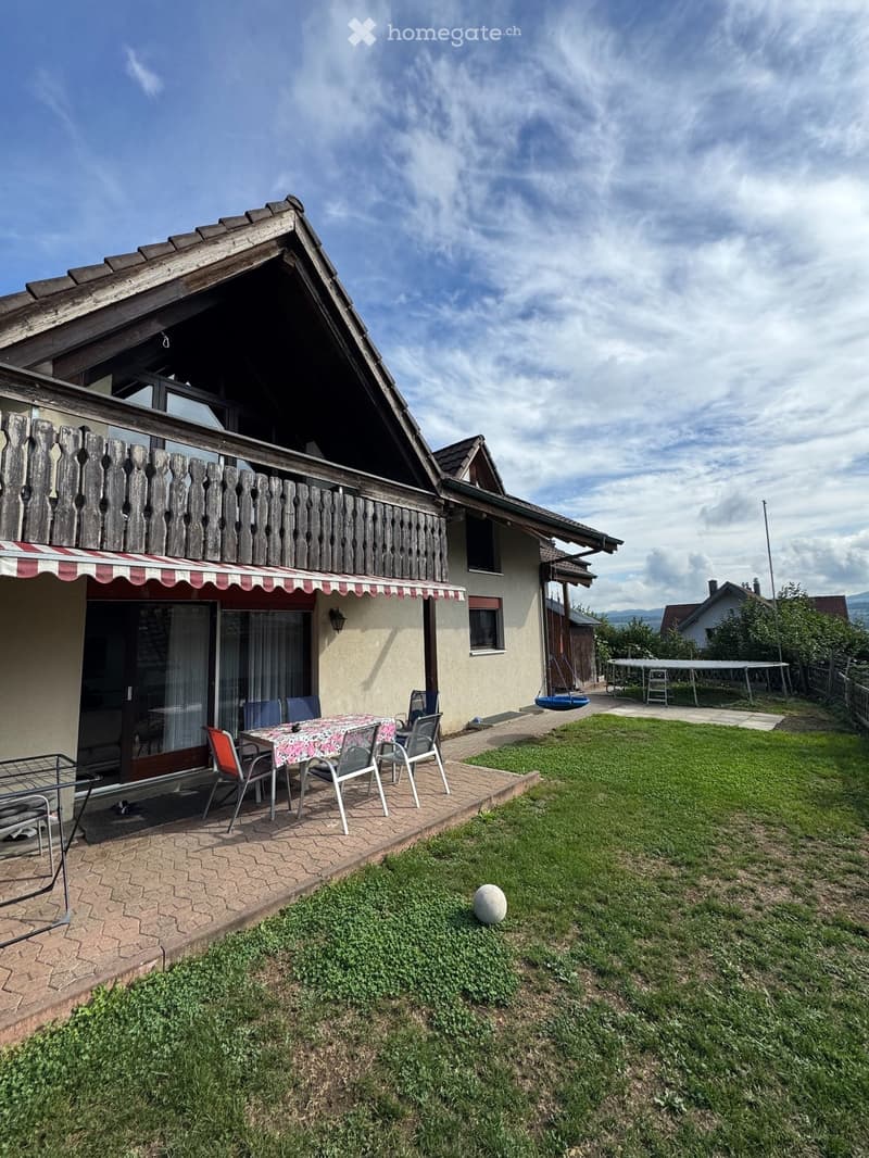 Garden area with a table, chairs, and a swing, adjacent to a house with a balcony and awning.