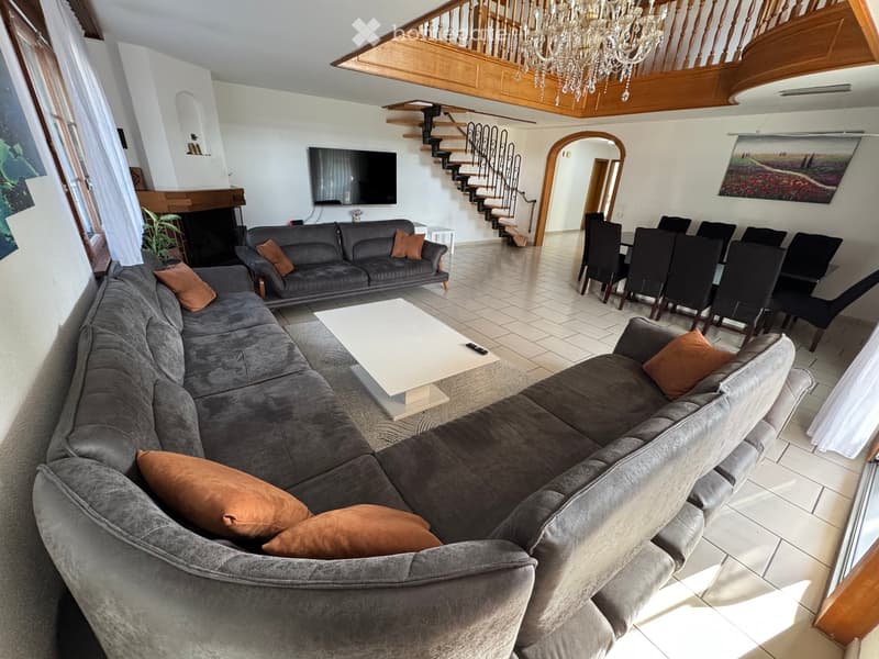 Spacious living and dining area with a large sectional sofa, coffee table, dining table with chairs, television, and a fireplace. The room features a staircase leading to an upper level and large windows with curtains.
