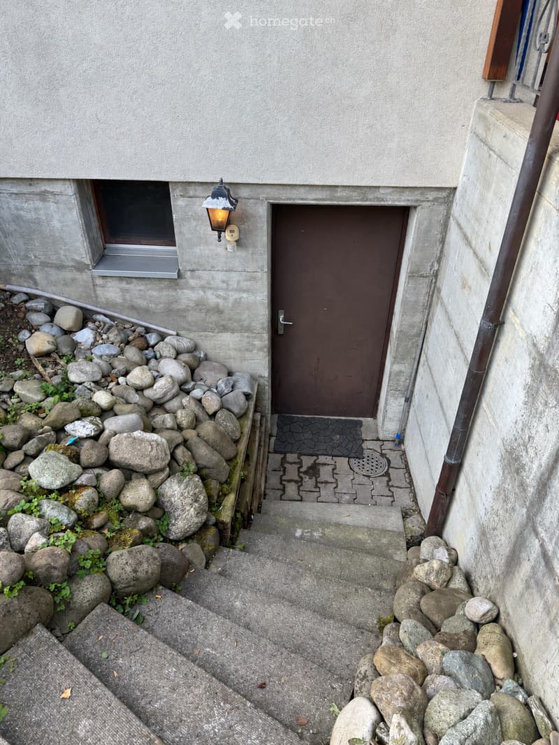 A narrow concrete staircase leading to a brown door, flanked by rock-filled planters and a small window on the left.