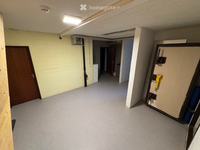 Empty hallway with concrete floor, exposed pipes, and a wooden door on the left. A small room is visible at the end of the hallway.