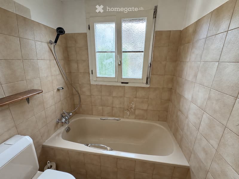 Bathroom with beige tiled walls and floor, a bathtub with a faucet, a toilet, a showerhead, and a wooden shelf above the bathtub.