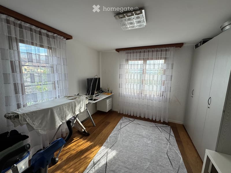 Well-lit room with wooden flooring, large windows with sheer curtains, an ironing board, a chair, a small table with a screen, and a cabinet.