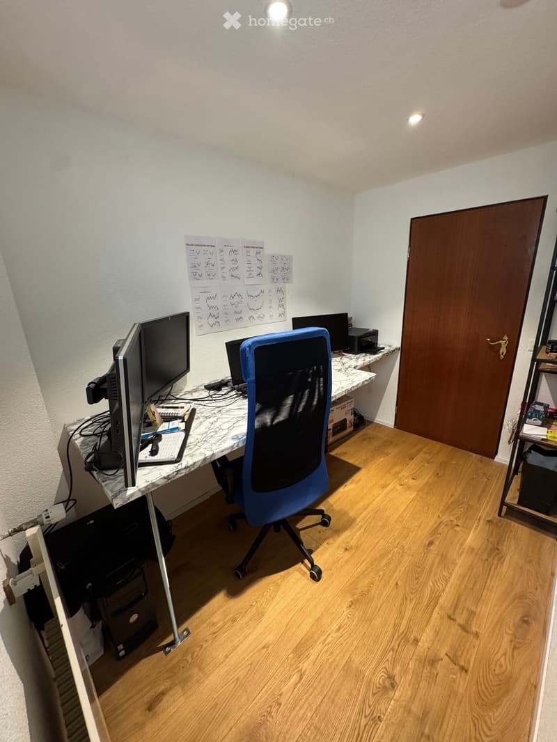 A well-lit room with wooden flooring, a white marble desk, a blue office chair, two computer monitors, a printer, and a shelf with various items.