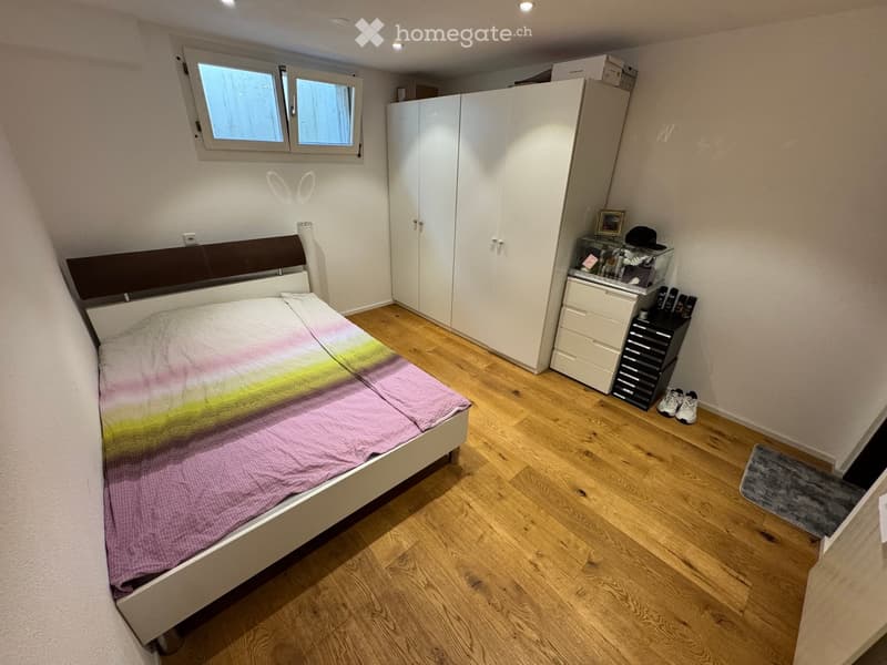 A modern bedroom with a wooden floor, a bed with a colorful bedspread, a large white wardrobe, a drawer unit, and a mirror. The room also includes a picture frame, a shoe rack, and a shower cap on the floor.