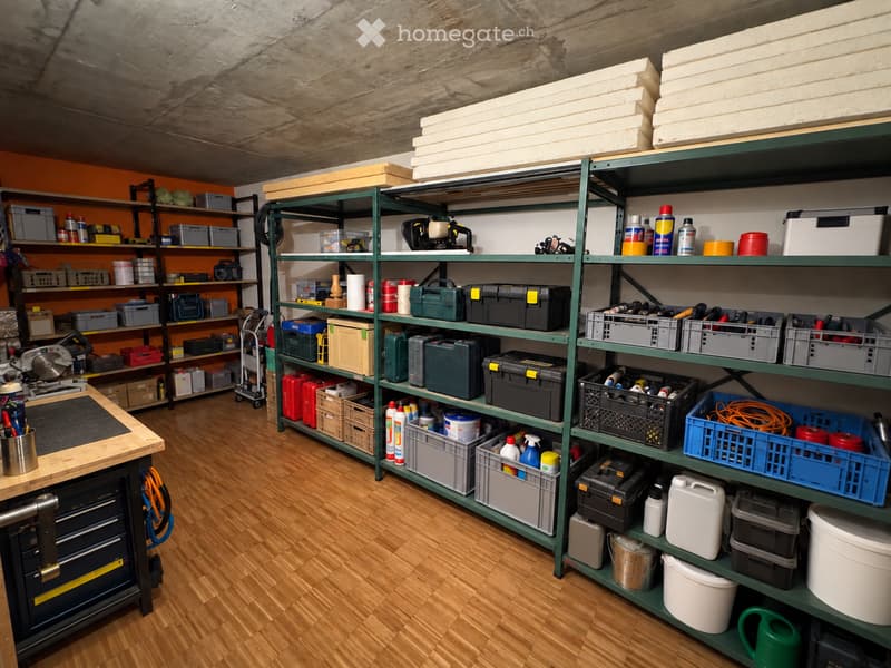 A well-organized workshop or storage room with wooden flooring, green metal shelving units filled with tools, boxes, and supplies, and a workbench with drawers.