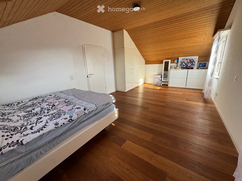 Bright bedroom with wooden floors, slanted wooden ceiling, bed with patterned bedding, white wardrobe, dresser, mirror, and picture frames on the wall.