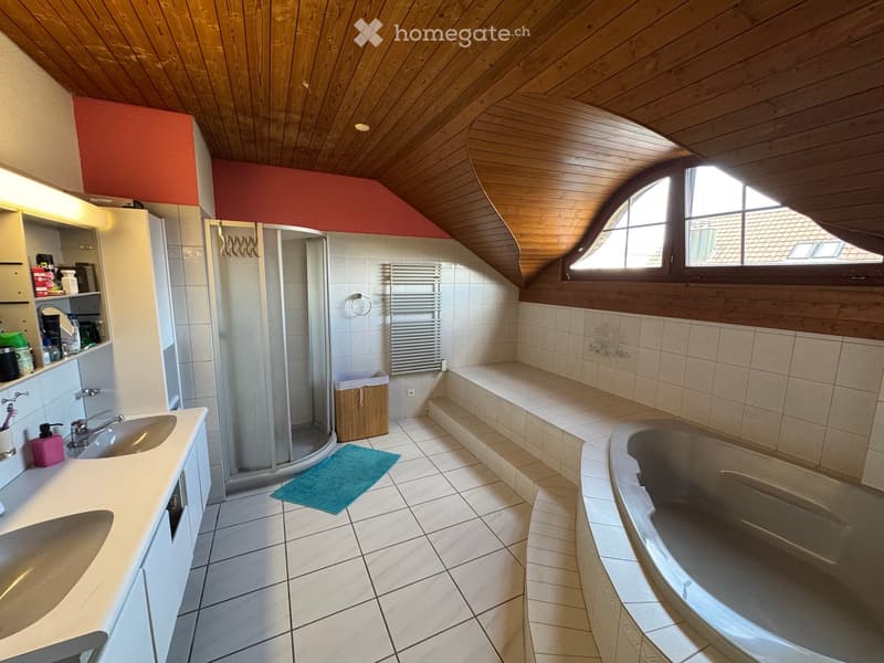 Bathroom with a wooden slanted ceiling, white tiled walls and floor, a large bathtub, a sink with a mirror, a glass-enclosed shower, and a radiator.