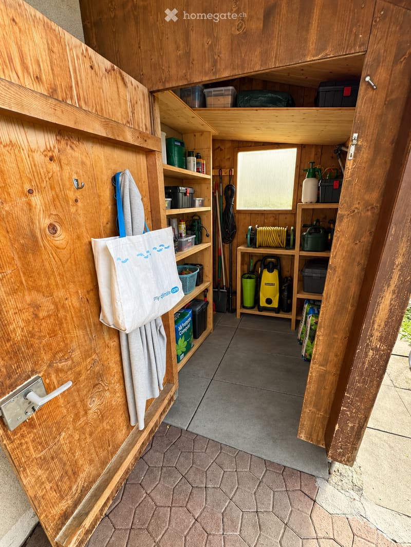 Wooden storage shed with shelves holding gardening tools, containers, and equipment. Features a tiled floor and a window for natural light.