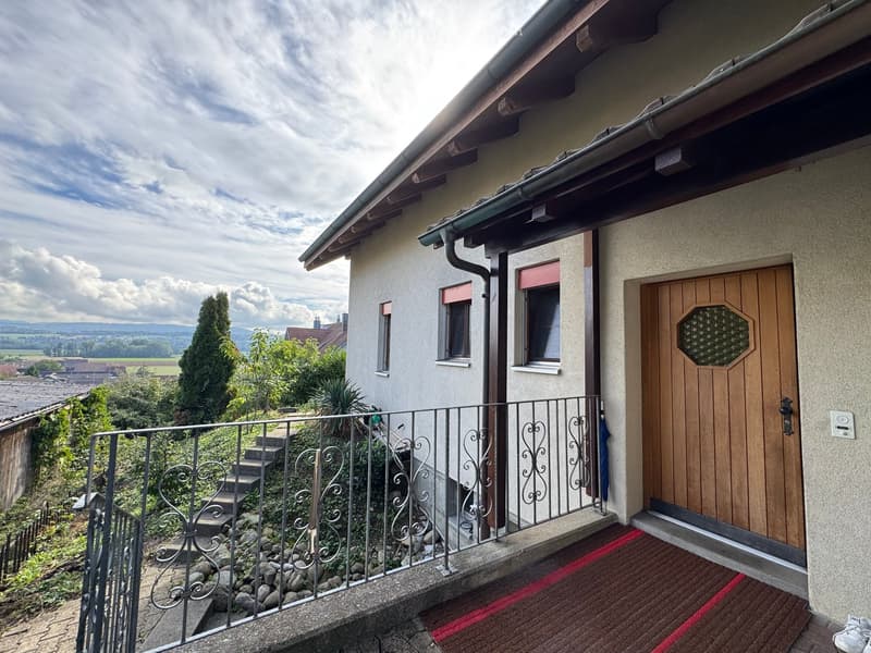 Balcony with metal railing, steps leading up, view of countryside and trees, wooden front door with decorative glass panel.
