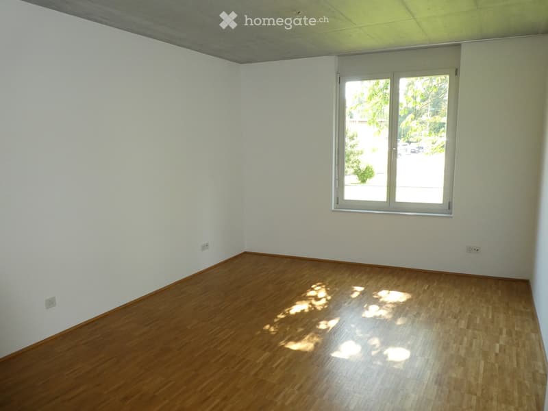 Spacious bedroom with hardwood flooring, white walls, and large windows providing natural light and a view of the surrounding greenery.