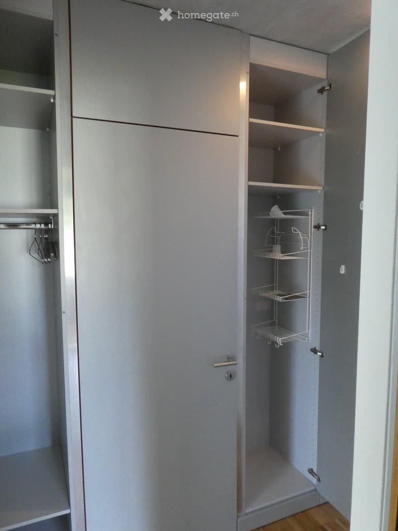 The image shows a bedroom closet with built-in shelves and hanging space. The closet has a gray, minimalist design with sliding doors. The interior of the closet has adjustable shelves and a hanging rod for clothes.