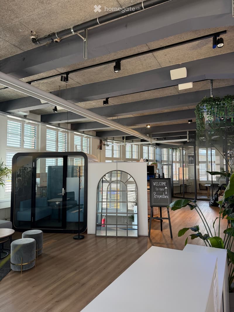 Modern office space with wooden flooring, large windows, and contemporary furniture including stools, tables, and a welcome sign.