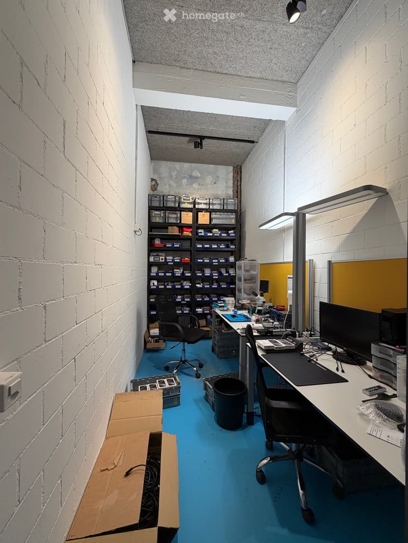 Office space with blue flooring, white brick walls, desks, chairs, shelves, and a microscope.