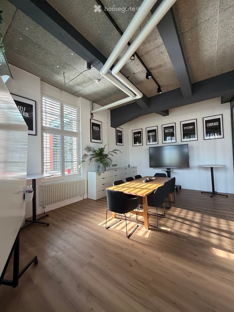 Modern office space with wooden flooring, large windows, a conference table with chairs, a TV screen, a cabinet, and framed motivational posters on the wall.
