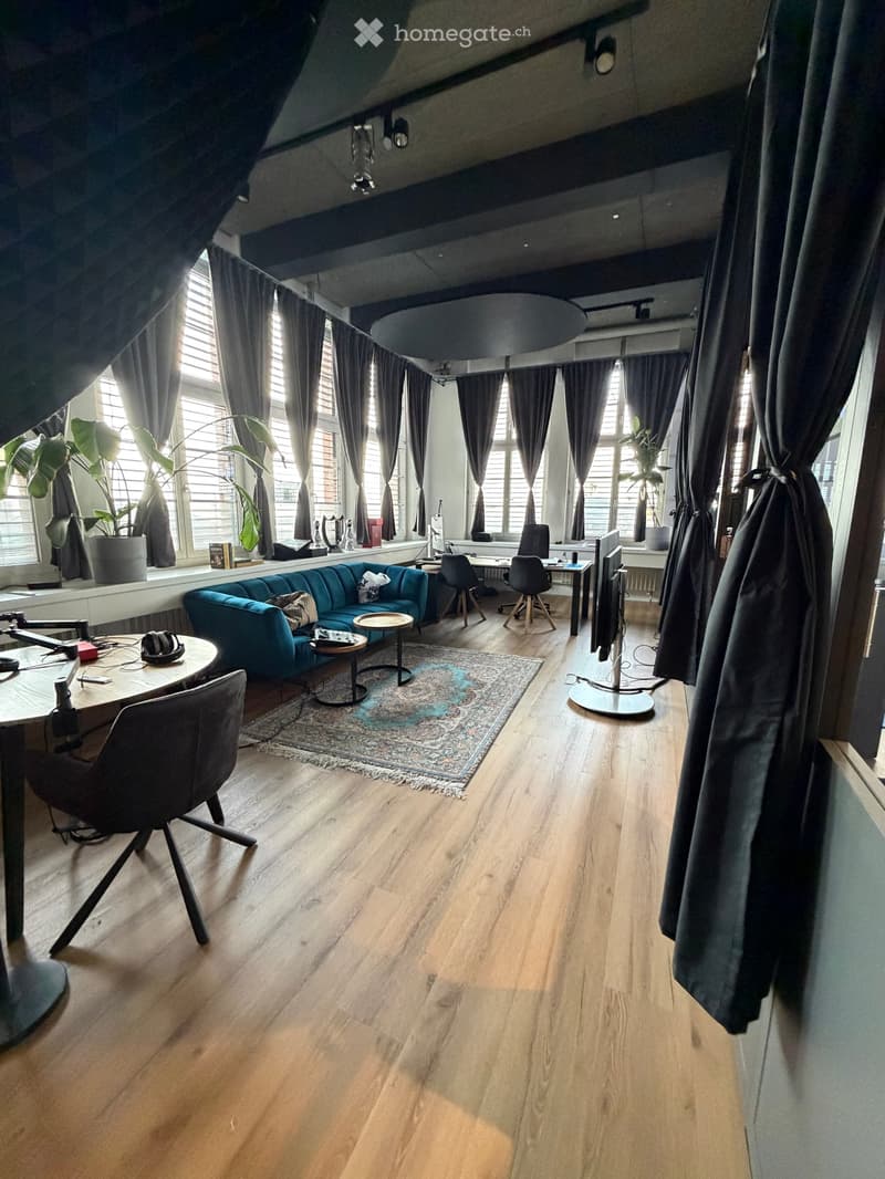 A modern, open-plan living and working space with wooden flooring, large windows with dark curtains, a blue sofa, a round coffee table, a patterned rug, a desk with chairs, and a large screen.