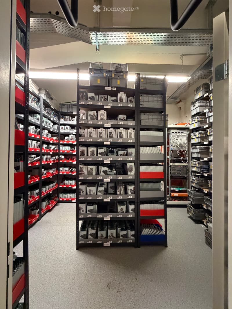 A storage corridor with multiple shelving units filled with books, boxes, and other items. The space has a carpeted floor and fluorescent lighting.