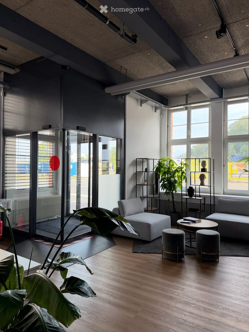 Modern living room with wooden flooring, large windows, grey sofa, round table, two stools, shelves, and plants.