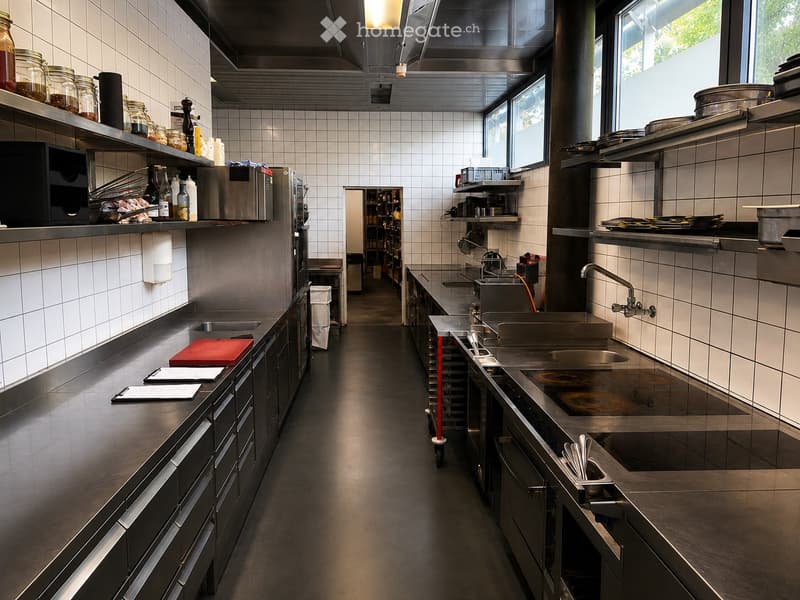 Commercial kitchen with stainless steel appliances, large windows, and organized shelves holding jars and bottles.