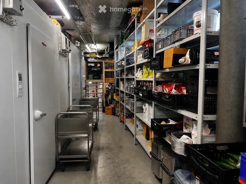 A well-lit storage corridor with metal shelving units filled with various items, including boxes, containers, and supplies. Stainless steel carts are positioned along the left side, and a large refrigerator is visible on the far left.