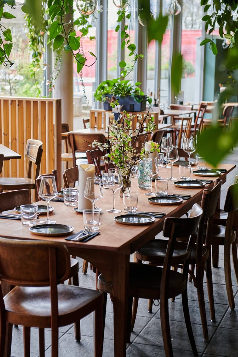 A restaurant setting with wooden tables and chairs, wine glasses, plates, cutlery, candles, and a flower arrangement on the table.