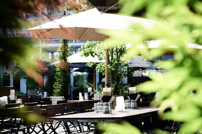 An outdoor dining area with tables, chairs, and umbrellas, surrounded by greenery and trees.
