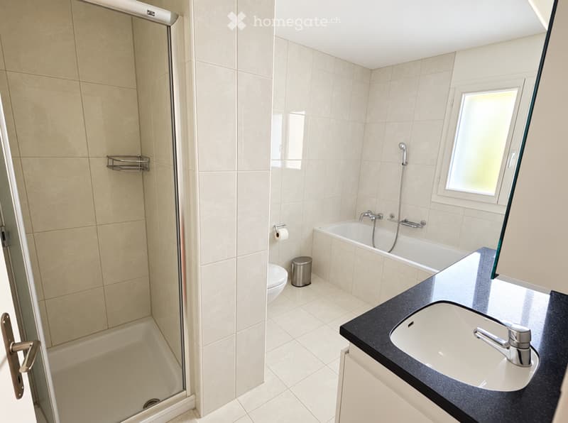 Modern bathroom with a bathtub, shower, sink, toilet, and a window providing natural light. The space features beige tiles and a sleek black countertop.